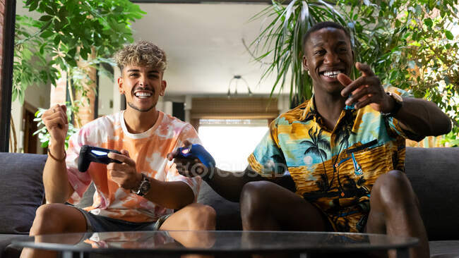 De baixo alegres amigos multirraciais com gamepads em mãos sentadas no sofá enquanto jogam videogame juntos na sala de estar leve com planta verde — Fotografia de Stock