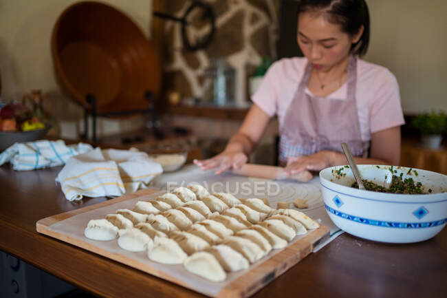 Сверху женщина в фартуке катит тесто на столе, пока готовит пельмени с мясом на кухне — стоковое фото
