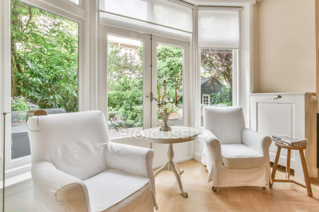 Salon contemporain intérieur avec chandelier sur la table entre fauteuils contre les fenêtres à la maison en journée — Photo de stock