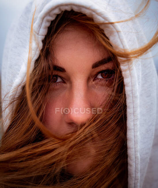 Portrait de charmante femme énigmatique aux cheveux roux volants et à la capuche regardant la caméra lors d'une journée venteuse en Galice — Photo de stock