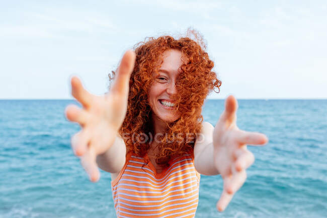 Optimistic young female with flying ginger hair reaching hands to camera on coast of blue rippling sea — Stock Photo
