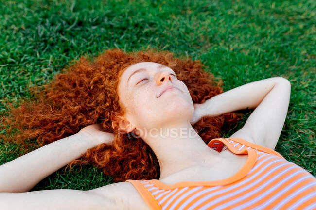Vista superior de la mujer de pelo rizado feliz con las manos detrás de la cabeza relajante en el césped con los ojos cerrados - foto de stock