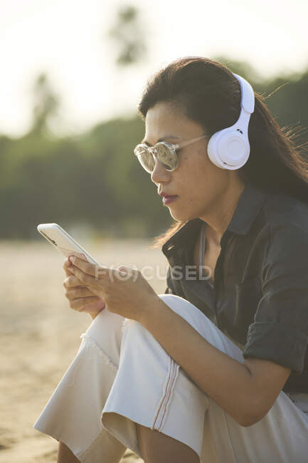 Vista lateral de mujer asiática seria en gafas de sol mensajería de texto en el teléfono celular mientras escucha música en auriculares en la orilla arenosa - foto de stock