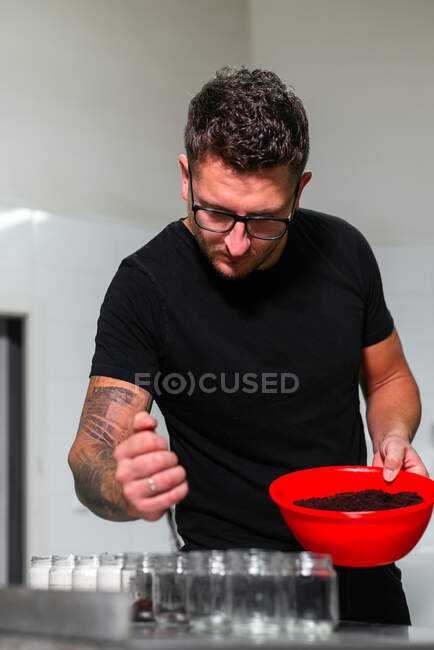 Focused young male baker in casual outfit and eyeglasses adding crumbs of chocolate cake into glass jars while preparing delicious layer dessert in kitchen — Stock Photo