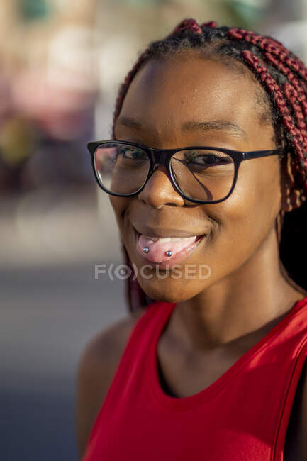 Позитивная афроамериканка с красными косичками в очках, показывающая язык с пирсингом и смотрящая в камеру на улице в солнечный день — стоковое фото