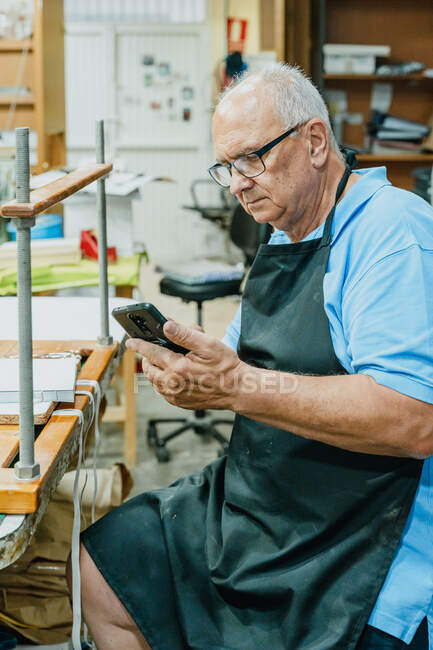 Concentrated senior male master in apron and eyeglasses using mobile phone while sitting at workbench during printing process in studio — Stock Photo
