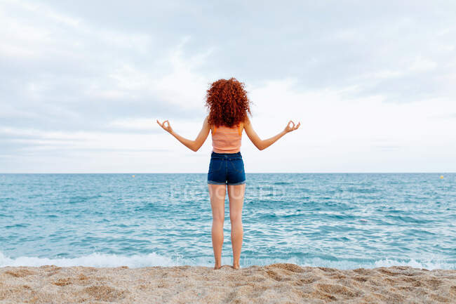 Обратный вид на неузнаваемую женщину с вьющимися рыжими волосами, делающими дзен-жест на берегу синего волнистого моря — стоковое фото