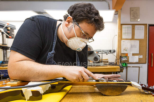 Side view of concentrated artisan in protective mask making carving on neck of guitar in workshop — Stock Photo