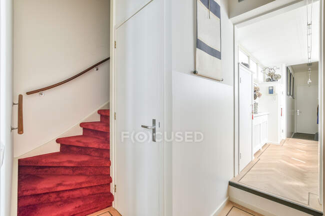 Красные ступеньки с деревянными перилами у белых дверей и стильная комната с висячими украшениями и шкафами в современной просторной квартире — стоковое фото