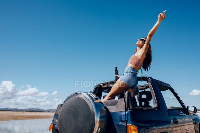 Full body of happy young female in summer outfit and sunglasses raising hand of beer while standing on roof of safari car on shore of river — Stock Photo