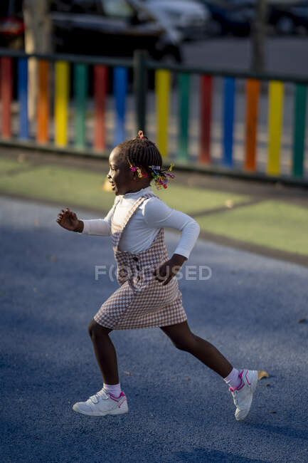 Vista lateral de la feliz chica afroamericana en vestido elegante y zapatillas de deporte corriendo en el patio de recreo a la luz del sol - foto de stock