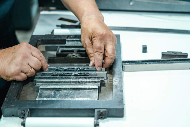 Crop unrecognizable elderly male artisan in apron and eyeglasses preparing metal frame for letterpress printing in studio — Stock Photo