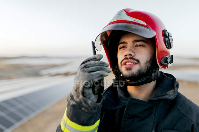 Positivo jovem bombeiro barbudo vestindo capacete e luvas e roupas usadas de proteção enquanto segurava walkie talkie na mão levantada e pacificamente olhando para o fundo turvo durante o dia — Fotografia de Stock