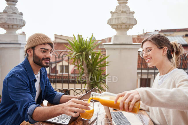Visão lateral da jovem fêmea positiva em óculos derramando suco fresco de garrafa de vidro em xícara de companheiro de apartamento masculino trabalhando no laptop na varanda — Fotografia de Stock