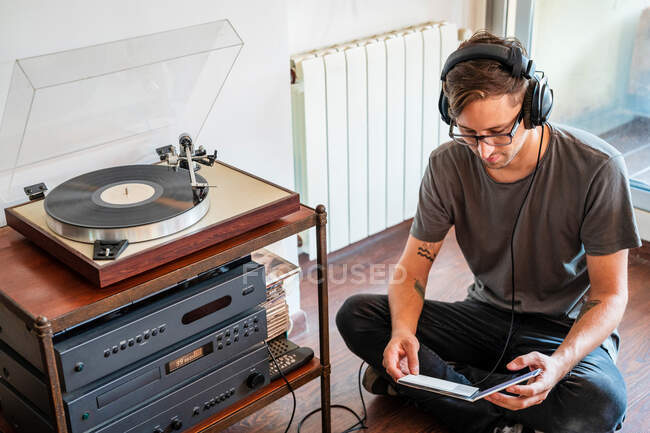 From above of man in casual clothes and headphones reading disc packaging while sitting on floor near record player — Stock Photo