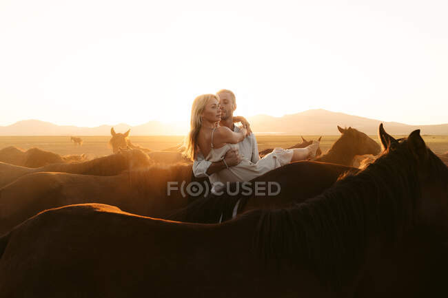 Homem segurando bela namorada de cabelos entre cavalos no campo pasto enquanto olha para longe — Fotografia de Stock
