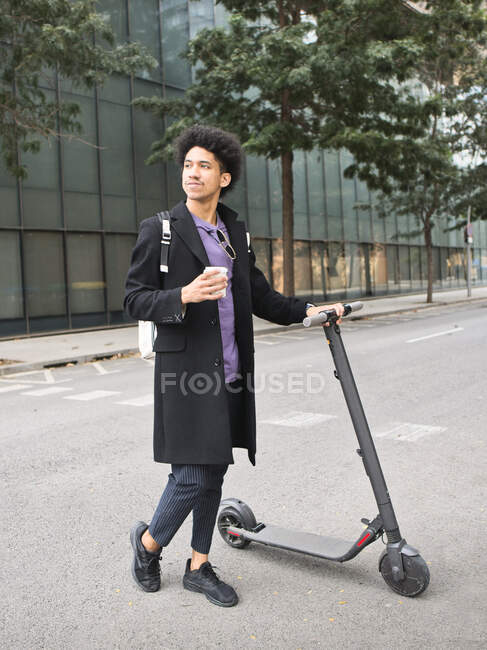 Joven y elegante hombre afroamericano con cabello oscuro rizado en elegante abrigo caminando por la calle con scooter eléctrico y una taza de café para llevar y mirando hacia otro lado - foto de stock