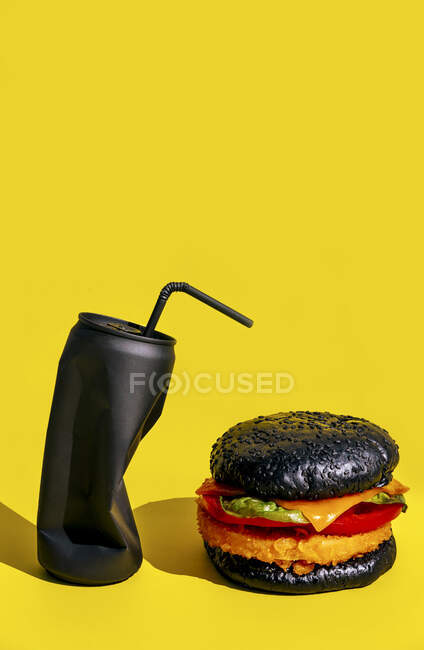 Hambúrguer fresco com legumes colocados perto de lata preta de refrigerante com palha contra fundo amarelo — Fotografia de Stock
