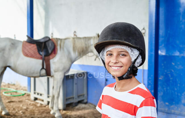 Niño sonriente con gorra de jockey y ropa casual mirando a la cámara mientras está de pie cerca del caballo blanco en el establo cerca de la pared del edificio a la luz del día - foto de stock