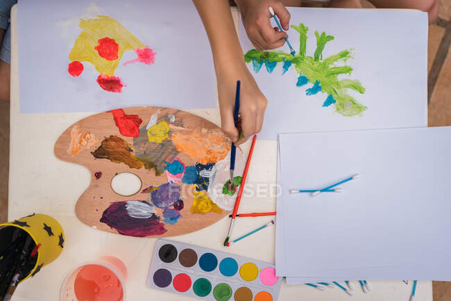 Dall'alto di coltura bambini anonimi che dipingono su carte bianche con pennelli a tavola con tavolozza di vernici in camera — Foto stock