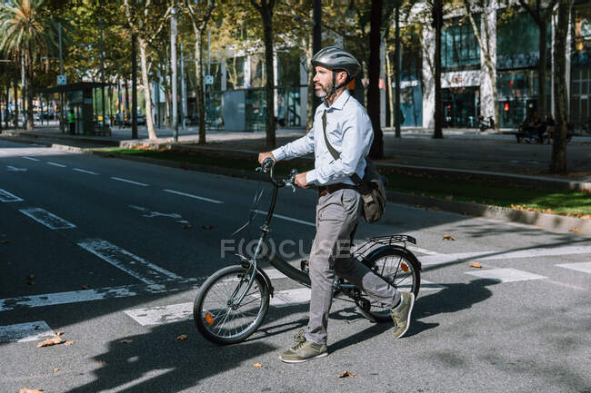 Toute la longueur du travailleur masculin barbu dans le casque traversant la route asphaltée avec vélo sur la route du travail — Photo de stock