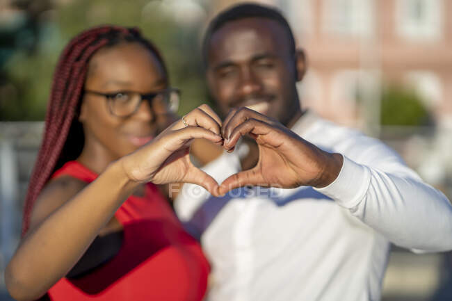 Feliz pareja afroamericana abrazando y mostrando un signo en forma de corazón con las manos mientras está de pie en la calle en un día soleado - foto de stock