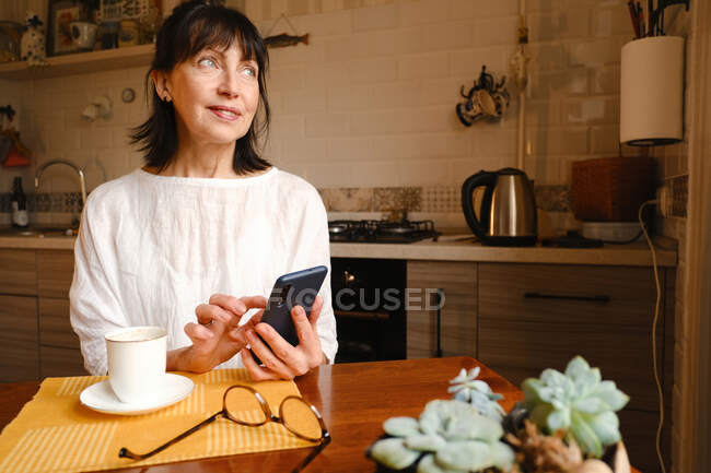 Satisfait textos féminins sur smartphone tout en étant assis à table avec une tasse de café sur la table dans la cuisine — Photo de stock