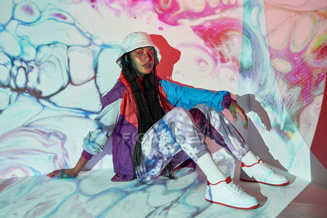 From above young Dominican female teenager in trendy outfit and hat sitting near white wall with creative abstract projections and looking at camera — Stock Photo