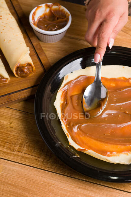 Crop faceless female spreading sweet Dulce de leche oh fresh crepe served on black plate on wooden table in kitchen — Stock Photo