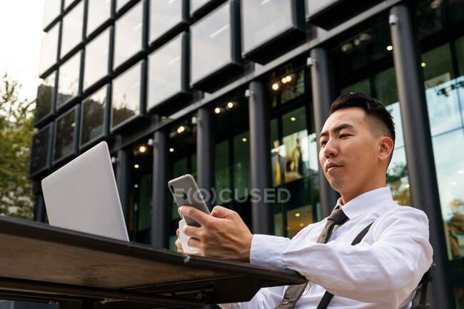 Desde abajo de joven empresario étnico masculino con café navegar por Internet en el teléfono celular mientras está sentado en la mesa de la cafetería urbana con el ordenador portátil - foto de stock