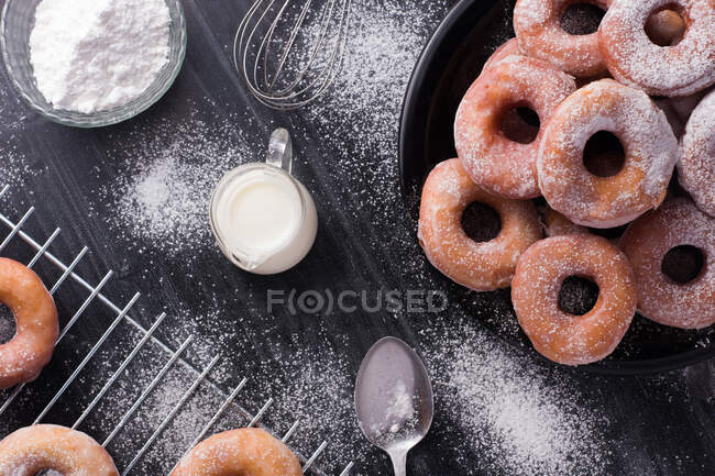 Doce frito donuts servido na placa perto de rack de refrigeração de metal e jarro de leite em mesa bagunçada preta com açúcar em pó — Fotografia de Stock