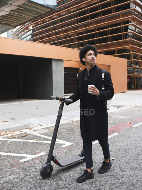 Joven y elegante hombre afroamericano con cabello oscuro rizado en elegante abrigo de pie en la calle con scooter eléctrico y una taza de café para llevar y mirando hacia otro lado - foto de stock