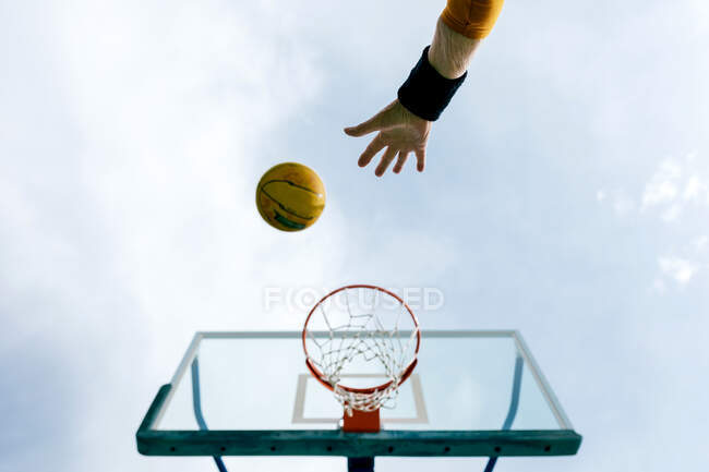 From below of crop anonymous person throwing basketball ball into hoop while playing game on public sports ground against blue sky — Stock Photo