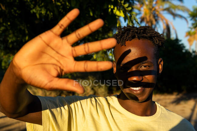 Улыбающийся молодой афроамериканец смотрит в камеру, закрывая лицо от солнечного света рукой в вечернее время. — стоковое фото