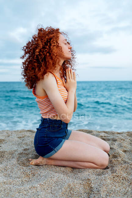 Side view full body of peaceful female with flying curly hair sitting on knees on sandy seashore and making namaste gesture during meditation — Stock Photo