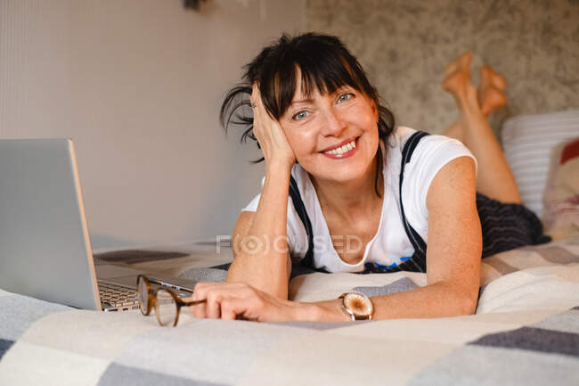 Full body of positive female leaning on hand while smiling brightly and chilling on bed near netbook with glance at camera — Stock Photo