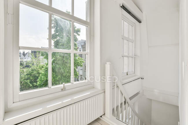 Chambre blanche vide avec escalier avec rampe près de la fenêtre en bois avec vue sur les bâtiments et les arbres verts — Photo de stock