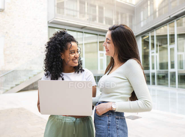 Cheerful multiracial coworkers in casual clothes standing with laptop on street and looking at each other against contemporary building with glass walls in daylight — Stock Photo