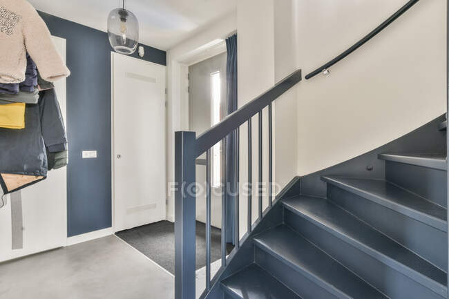 Scala blu con ringhiera situata vicino a porte e capispalla nel corridoio illuminato dal sole della casa moderna — Foto stock
