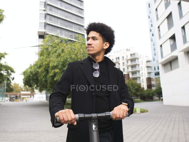 Нахабний молодий афроамериканець з темним кучерявим волоссям в стильному спорядженні стоїть на міській площі і озирається назад після їзди на скутері — стокове фото