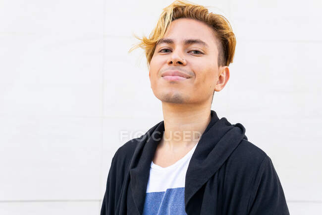 Young male in modern black cardigan looking at camera with thoughtful glance while standing near white wall on street — Stock Photo