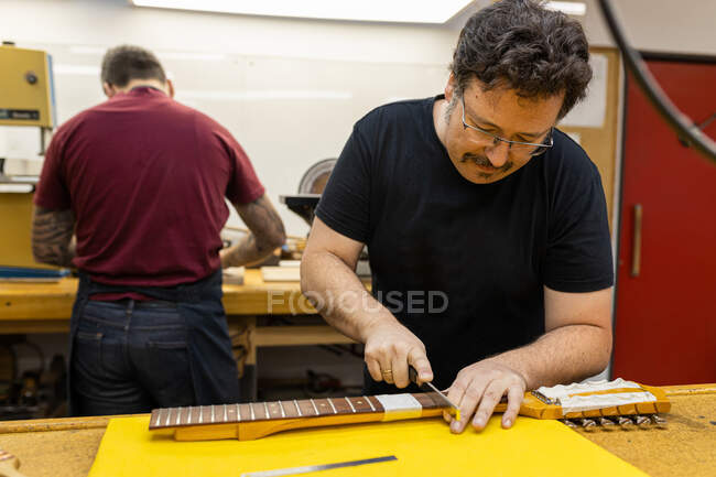 Maître de guitare concentré debout à table et couronnant des frets sur le cou de la guitare pendant le travail avec un collègue dans un atelier professionnel — Photo de stock