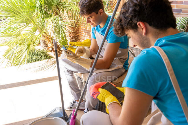 Full body of young male professional cleaning workers in uniform and gloves using mobile phones while resting on cozy couch on terrace of modern cottage — Stock Photo