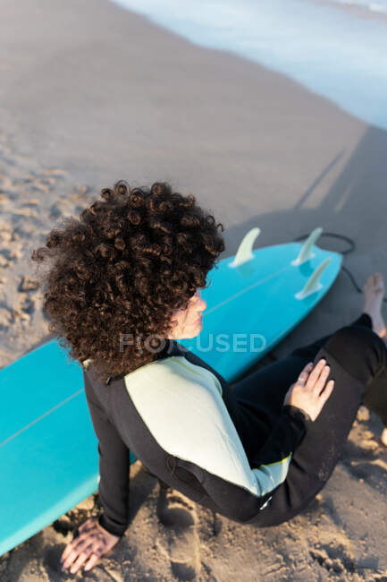 From above female surfer in wetsuit sitting on sandy beach near waving sea looking away — Stock Photo