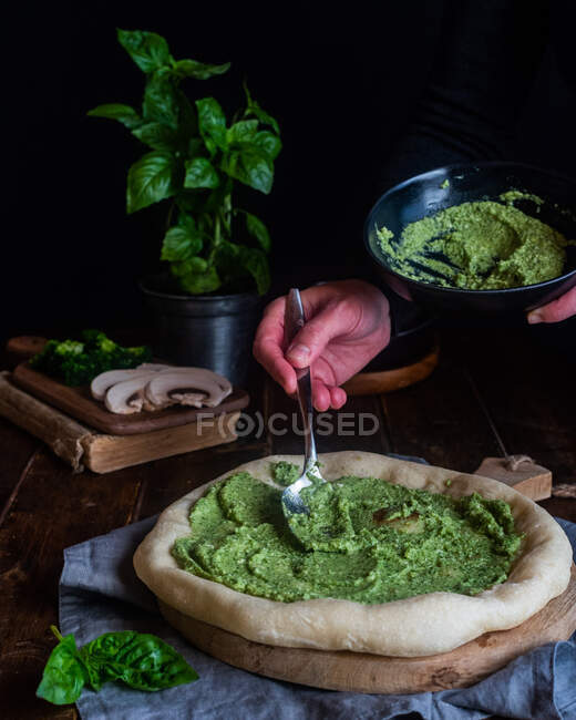 Cultivo irreconocible chef añadiendo salsa de pesto verde y cocinar deliciosa pizza vegetariana en la mesa oscura - foto de stock