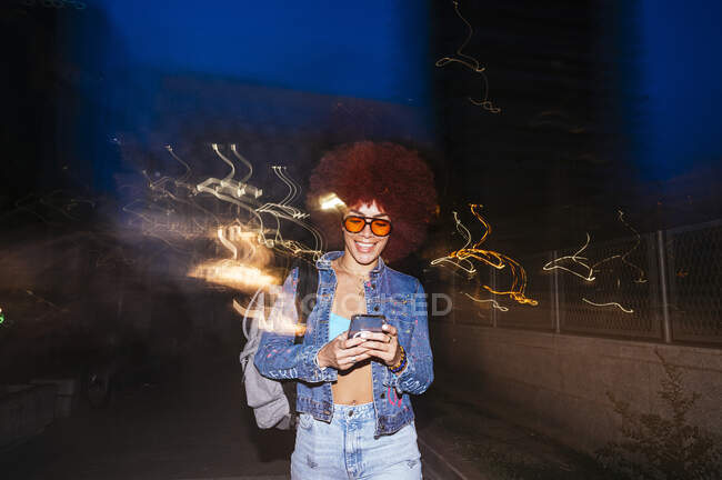 Mulher alegre com óculos de sol enviando mensagem de texto enquanto está perto da cerca com efeito de luz à noite — Fotografia de Stock