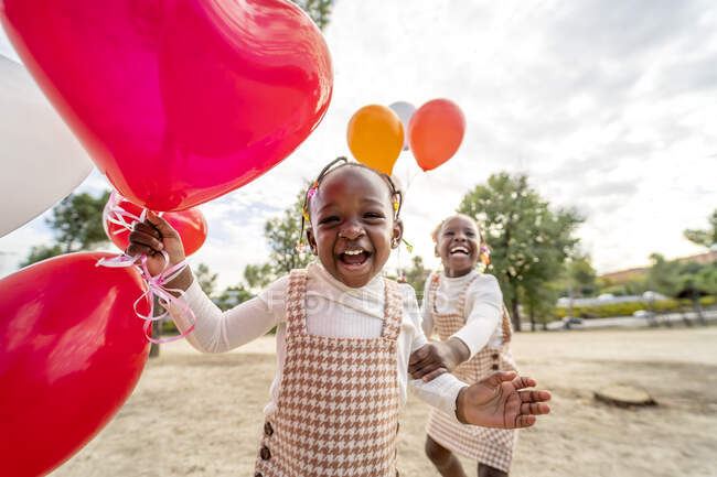 Fröhliche afroamerikanische kleine Schwestern in ähnlichen Kleidern stehen mit bunten Luftballons in der Hand auf grünem Gras im Park bei Tageslicht — Stockfoto