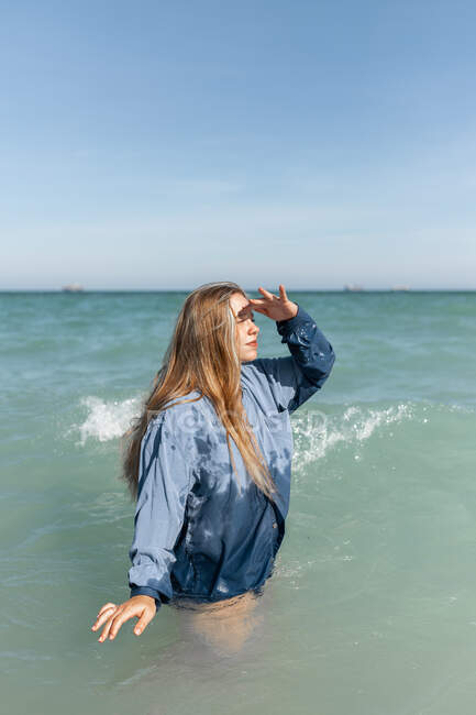 Side view of charming young female in wet shirt standing in sea water and looking into distance in sunny summer day — Stock Photo