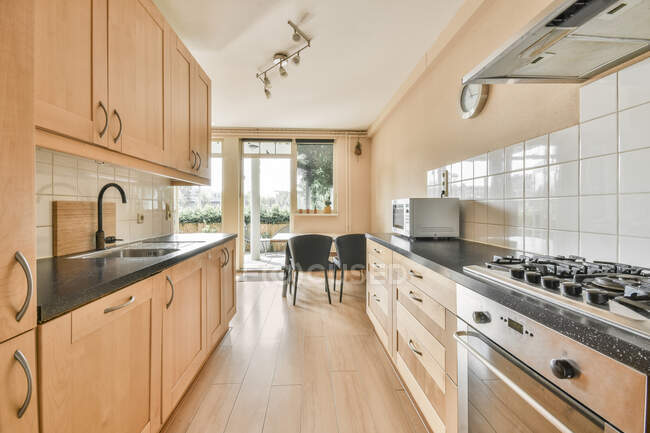 Diseño interior contemporáneo de cocina moderna amueblada con estantes de madera y zona de comedor equipada con electrodomésticos a la luz del día - foto de stock