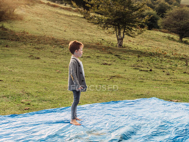 Seitenansicht des niedlichen barfüßigen kleinen Jungen in stylischer Kleidung, der auf einem Zelt steht und am grasbewachsenen Berghang des Campingplatzes wegschaut — Stockfoto